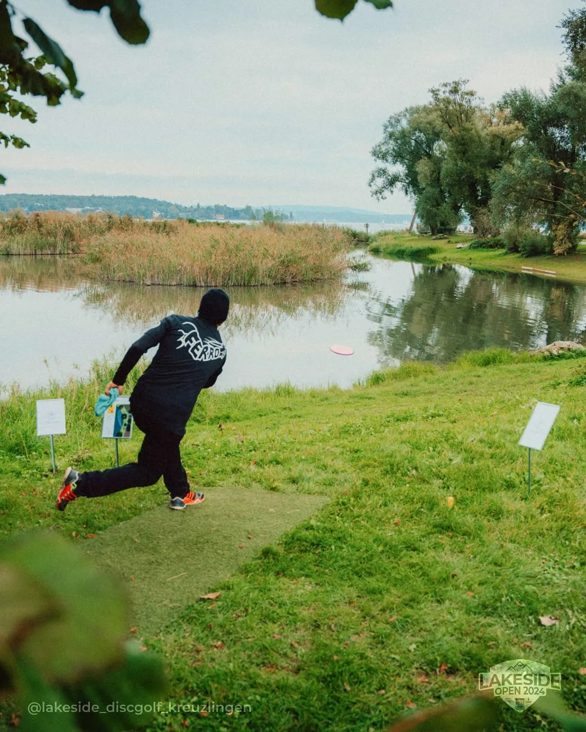 Spieler beim Abwurf über einen Weiher mit Schilf, im Hintergrund der Bodensee