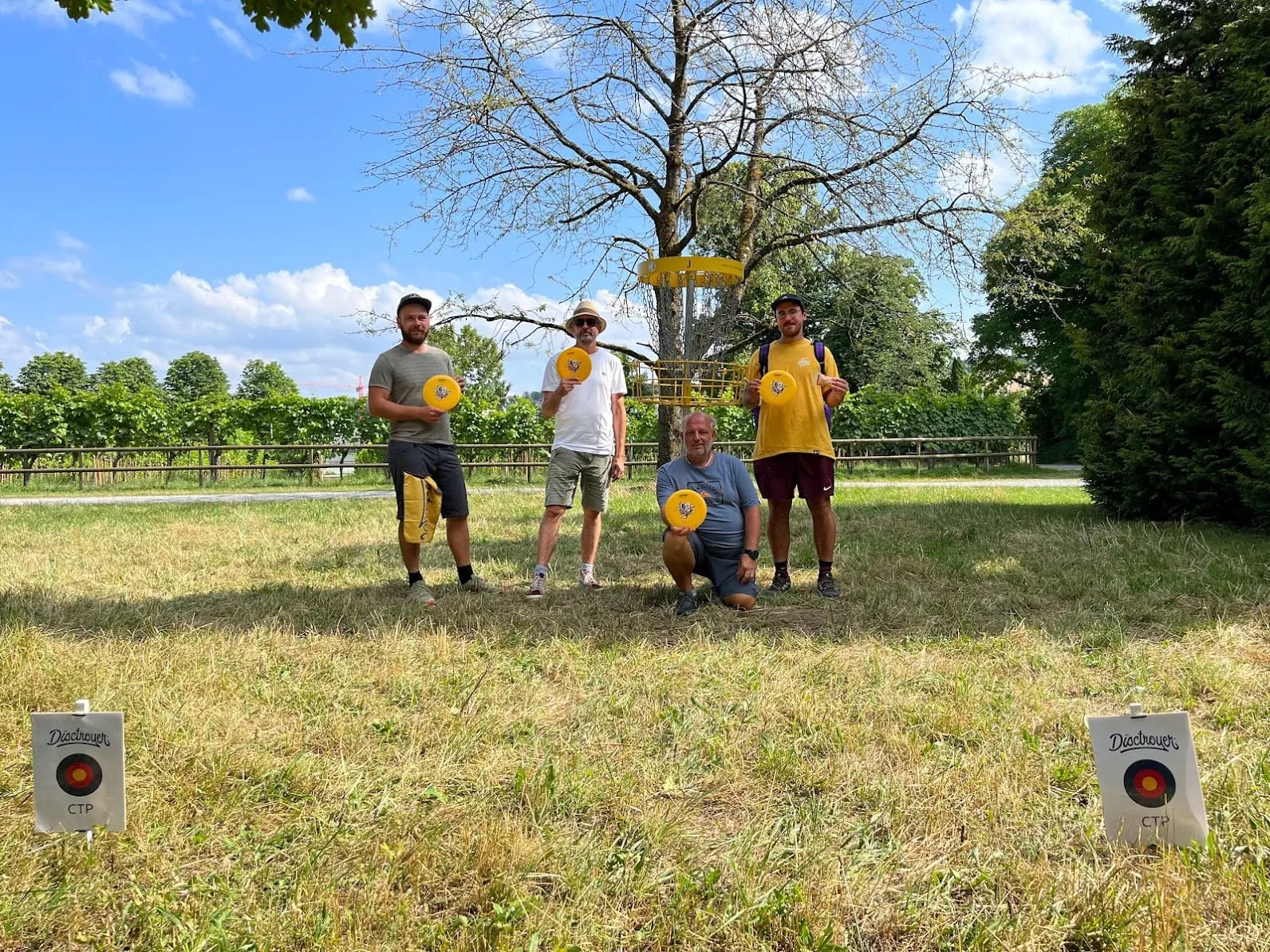 Vier Spieler stehen mit Discs unter einem Baum mit Disc Golf Korb