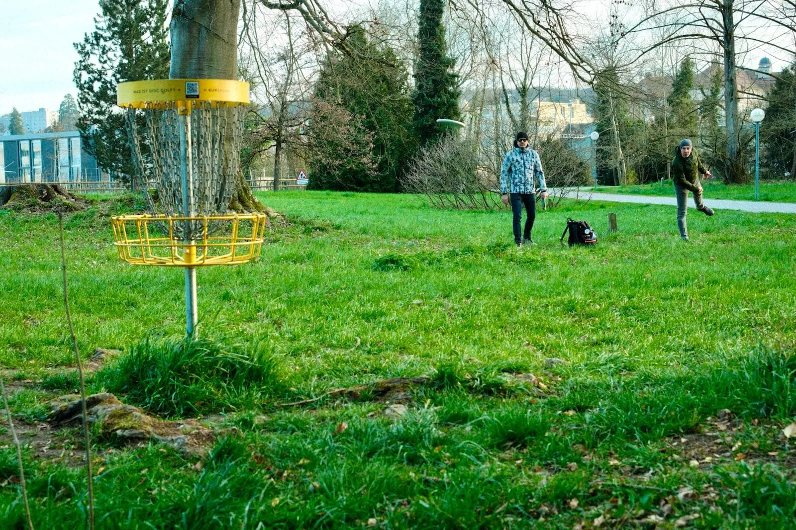 Gelber Disc Golf Korb auf der Wiese im Seeburgpark, im Hintergrund zwei Spieler